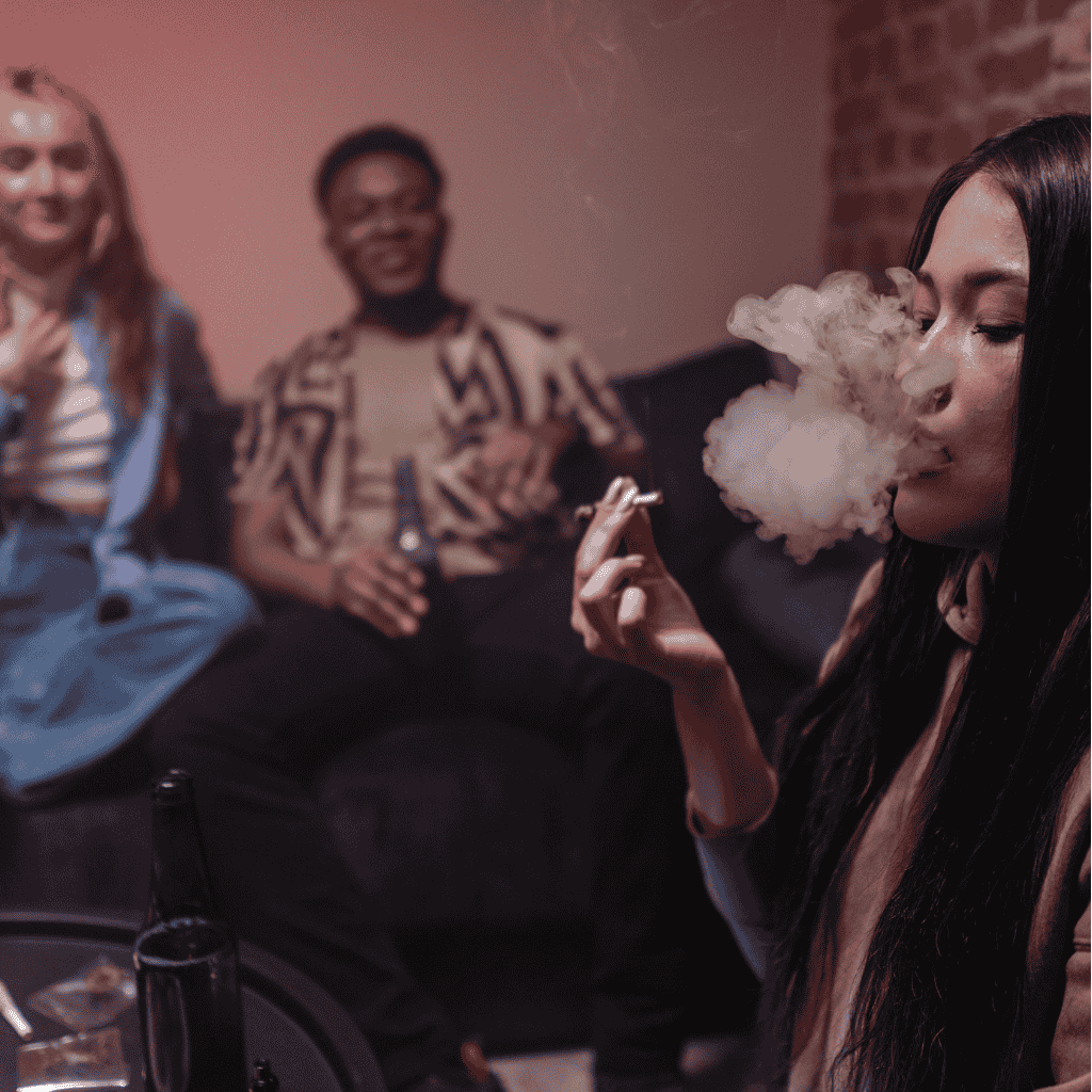 A woman in the foreground with smoke in front of her mouth. In the background are 2 more people